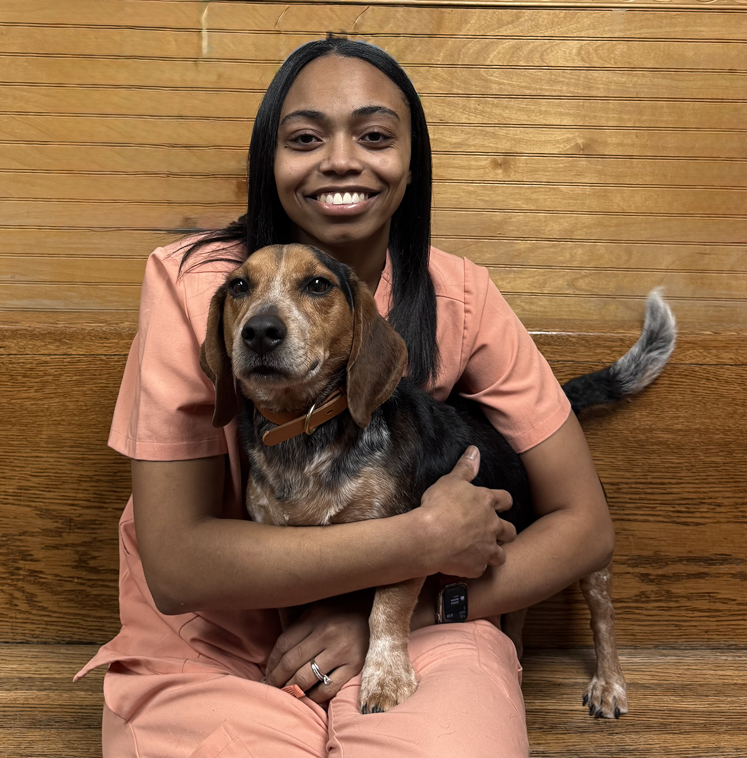 A smiling woman in pink scrubs hugging a beagle dog indoors.